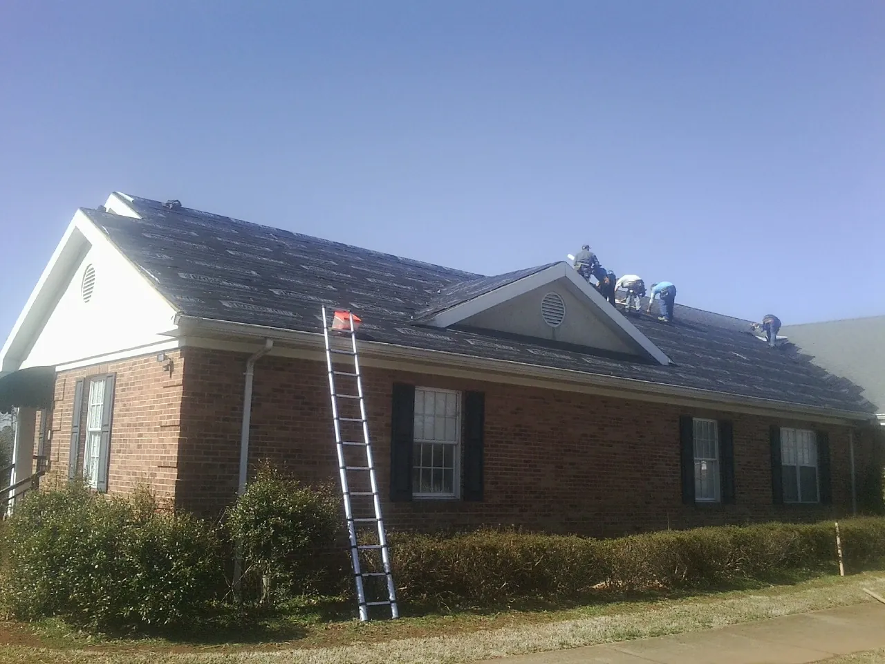 Crew tearing shingles off a brick ranch roof with a ladder set — Metal Roof Repair work by our crew in Blandinsville