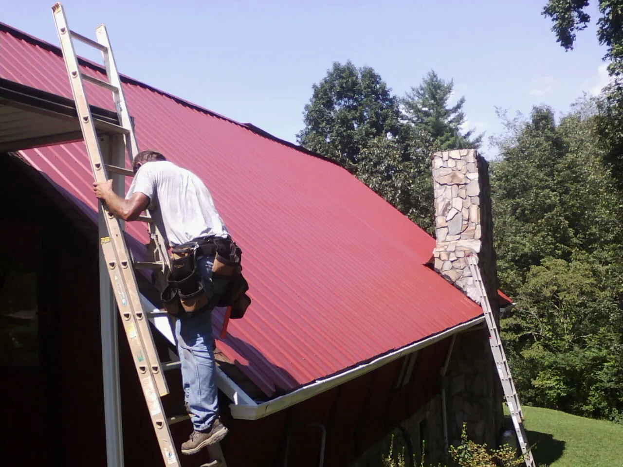 Roofer on a ladder beside a red metal roof and stone chimney — Soffit & Fascia Repair by Roofing Brothers in Blandinsville